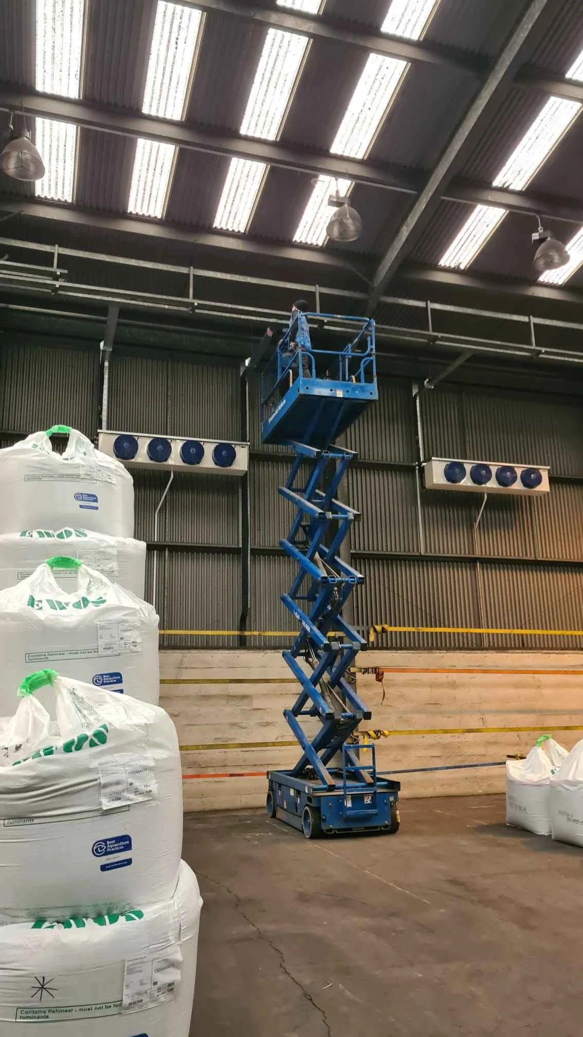 Person using a blue scissor lift inside a warehouse, surrounded by large white bags and industrial lighting.