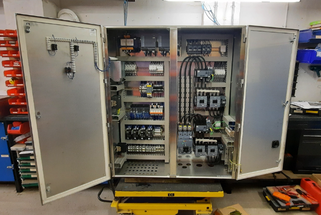 Open electrical control panel with various circuit breakers, relays, and wiring inside, mounted on a workbench in an industrial or maintenance setting.