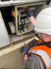 Electrician working on an outdoor electrical panel with wires and circuit components, wearing a hard hat and safety gloves.