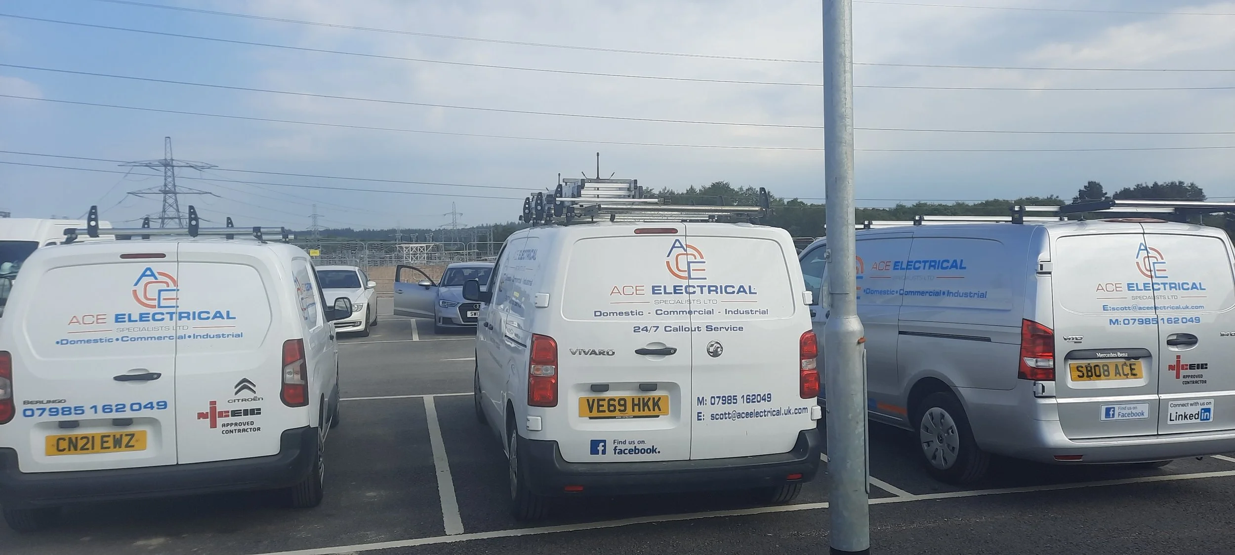 Parking lot with multiple white service vans labeled 'ACE Electrical,' parked next to a silver vehicle and some cars in the background, with power lines and a cloudy sky overhead.