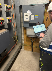 A man holding a laptop in front of electrical equipment and control panel in a utility or industrial setting.