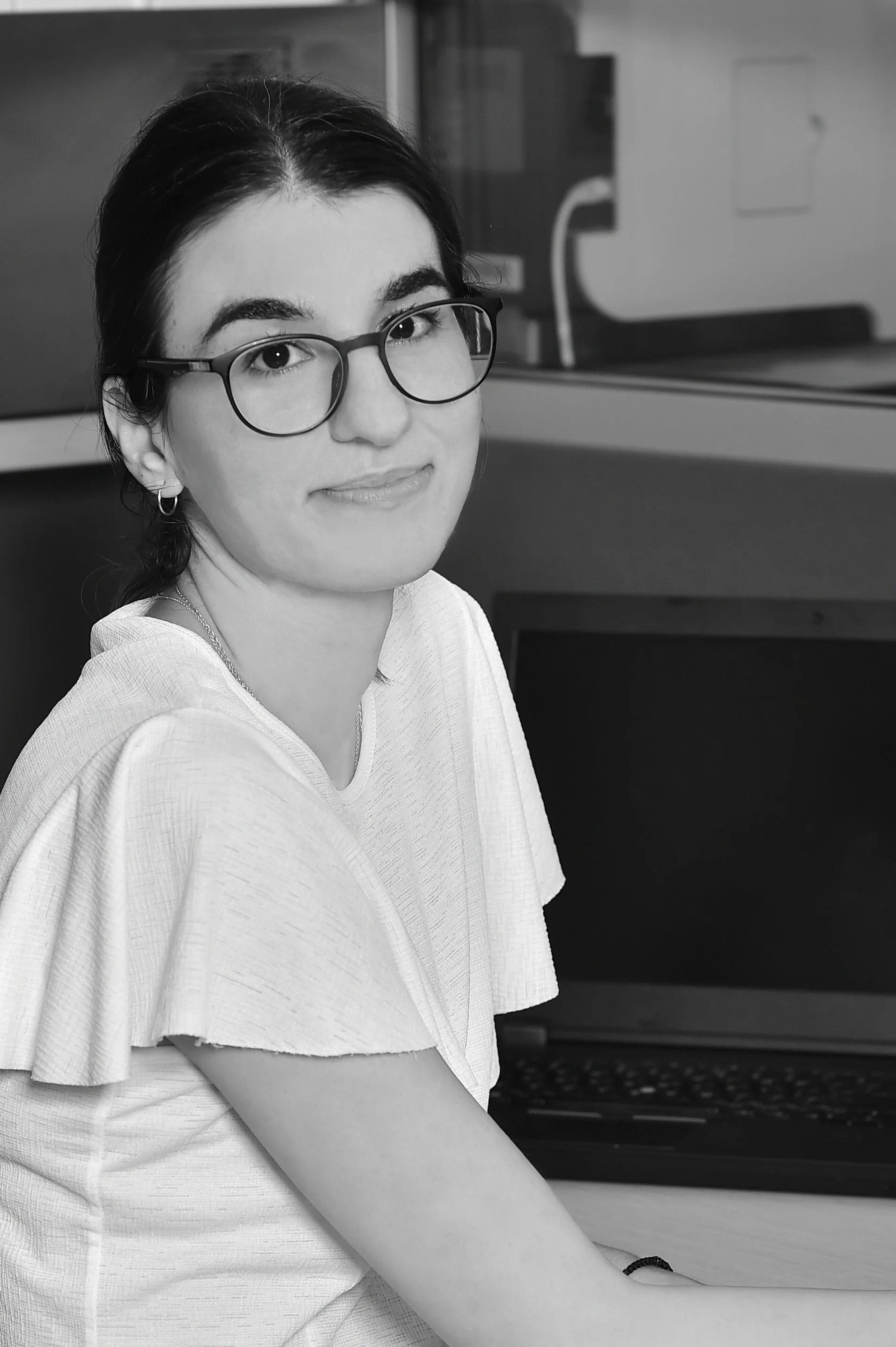 A woman with dark hair wearing glasses and a white top, sitting at a desk with a laptop, looking at the camera.