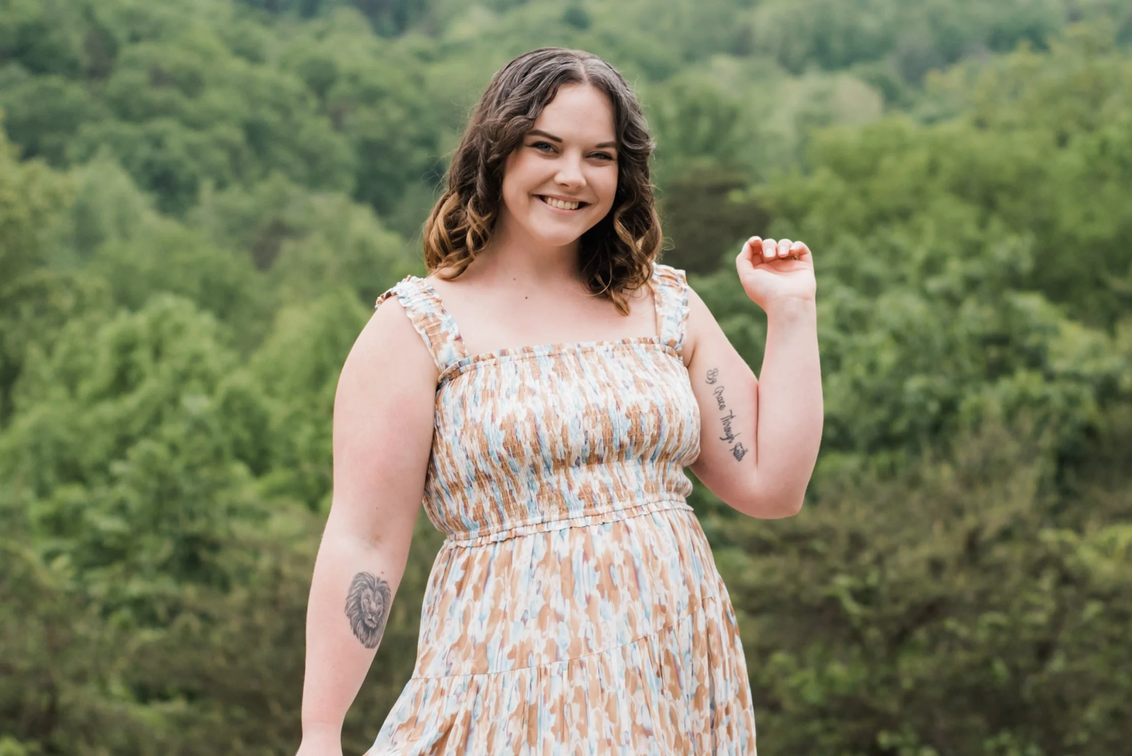 A smiling woman with shoulder-length wavy brown hair, wearing a sleeveless patterned sundress, stands outdoors with a background of green trees. She is raising her right arm with a fist and has tattoos on her left arm, including a lion's face and script text.