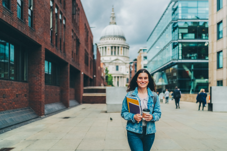 Young woman smiling and holding books and a coffee cup on a city street with the U.S. Capitol building in the background.