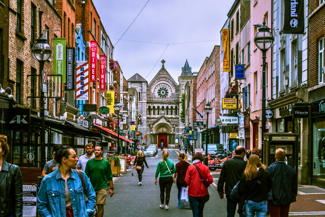 A busy city street in Dublin with pedestrians walking past shops and signs, leading to a historic church in the background.