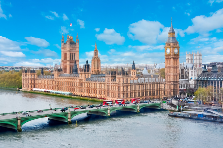 The Palace of Westminster, including Big Ben, viewed from the River Thames with Westminster Bridge in the foreground and a partly cloudy sky.