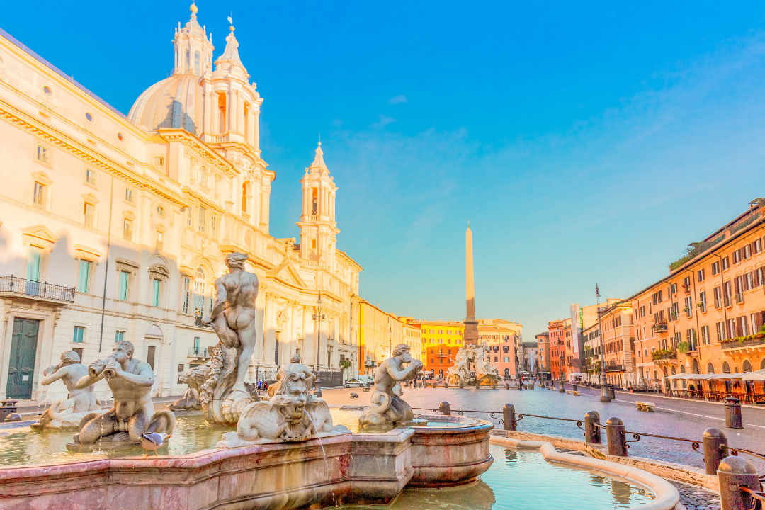 A city square with a large historic church and fountain with statues in the foreground, under a clear blue sky.