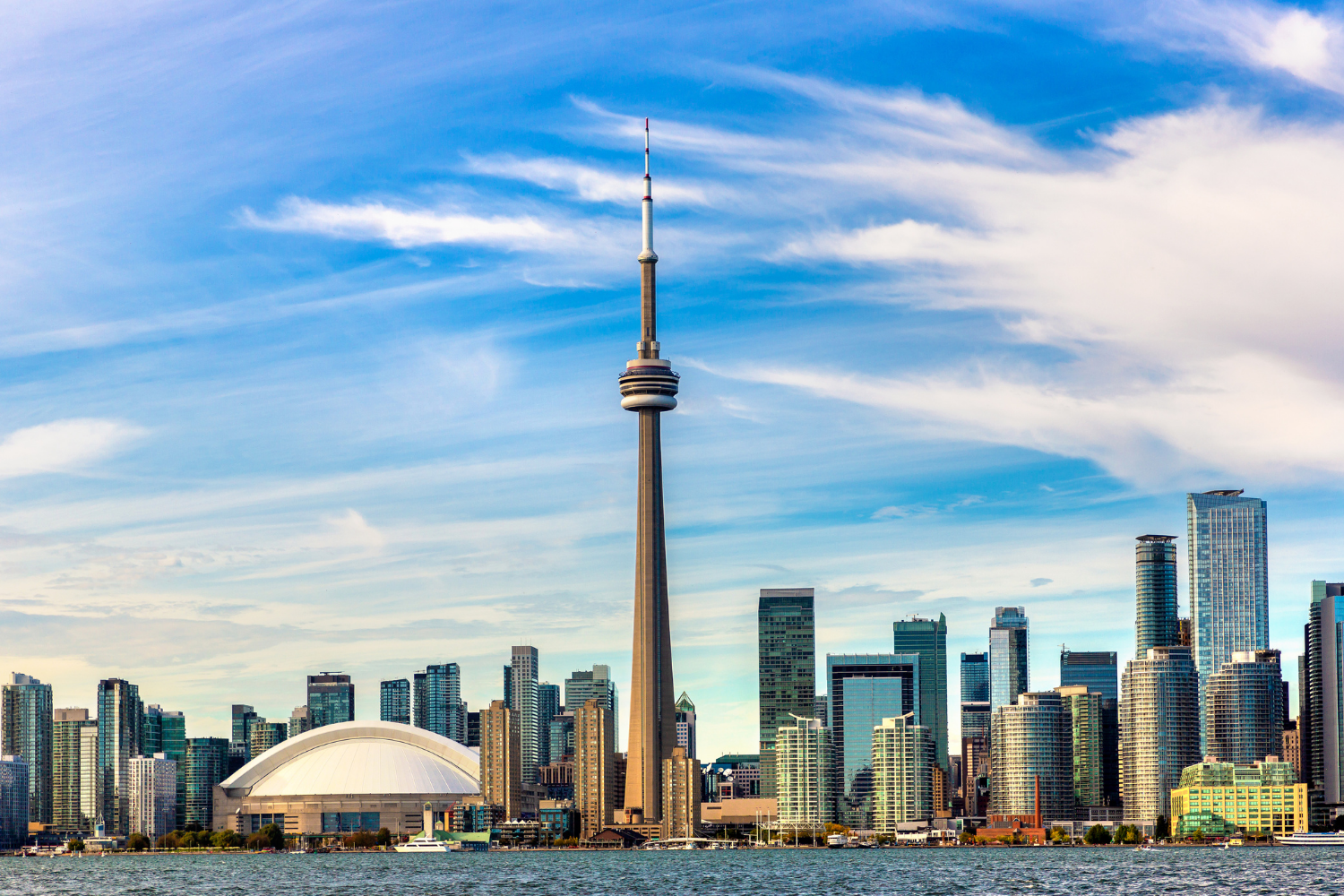 Skyline of downtown Toronto, Canada, featuring the CN Tower, Rogers Centre stadium, and modern skyscrapers along Lake Ontario with a partly cloudy sky.