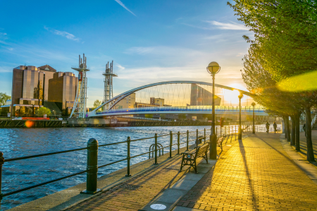 A riverside walkway at sunset with benches, trees, and a view of a bridge and buildings in the distance.