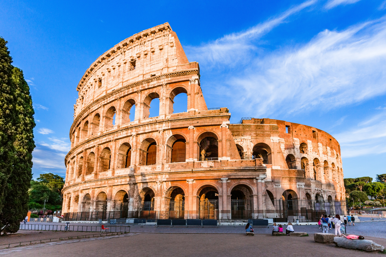 The Colosseum in Rome, Italy, with a clear blue sky, surrounded by visitors sitting and walking around.