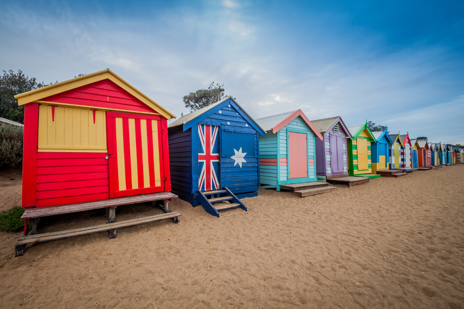 Colorful beach huts lined up on sandy beach, under a cloudy sky.