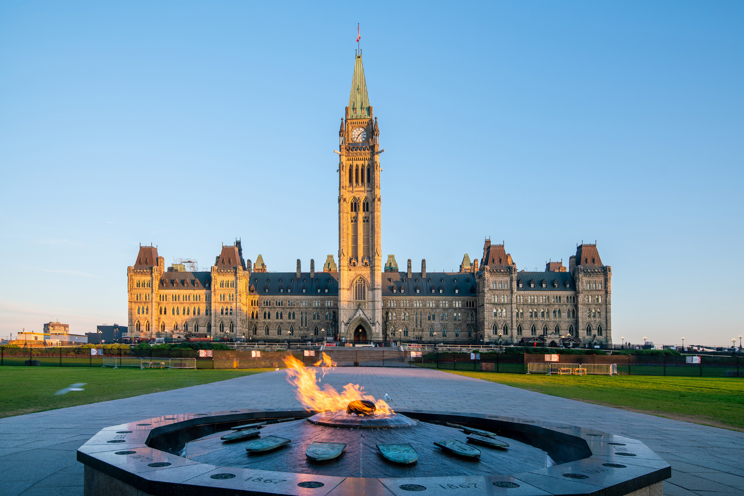 Front view of the Parliament Hill in Ottawa, Canada, with a memorial flame in the foreground and a clear blue sky.