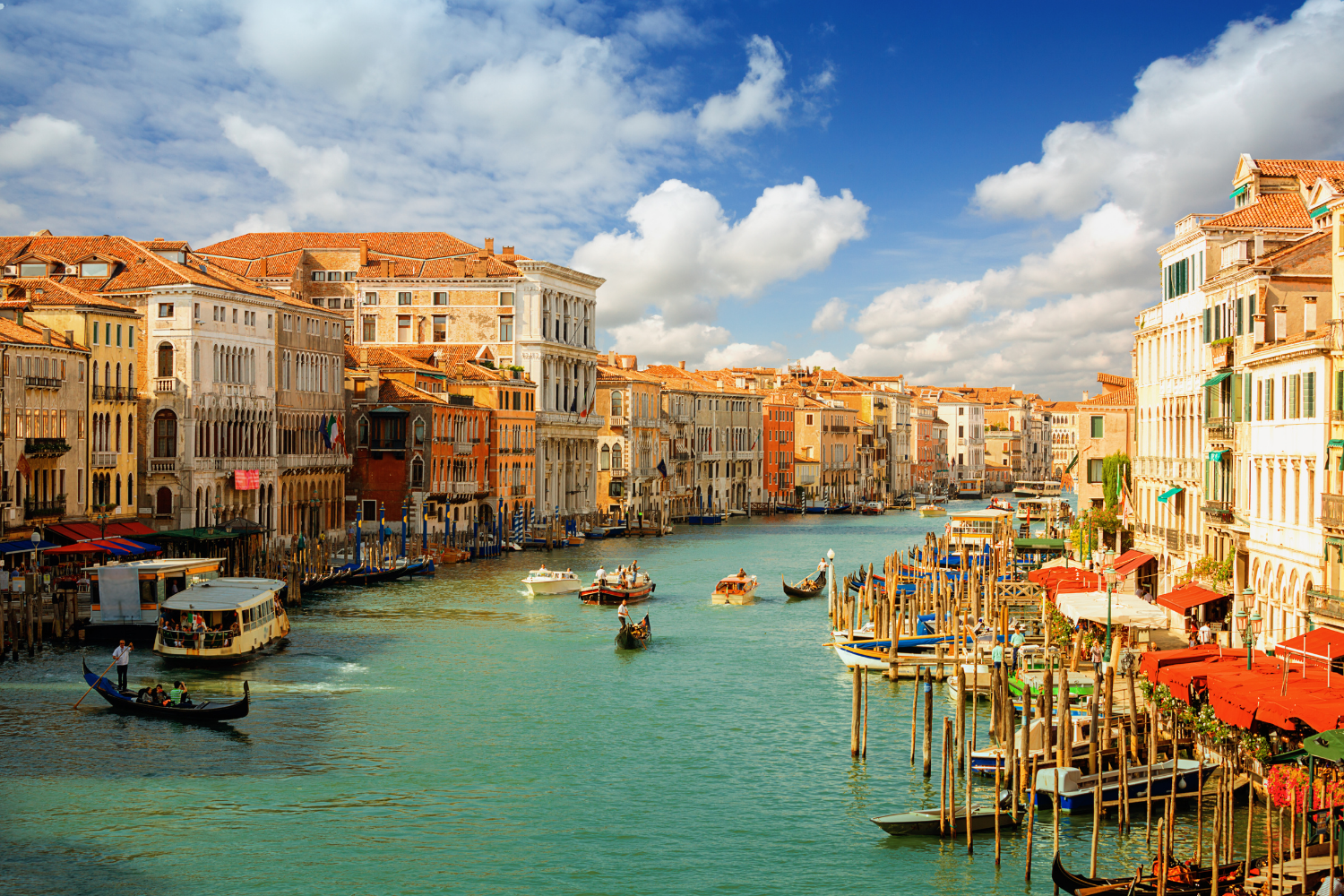 View of a canal in Venice, Italy, with boats, gondolas, and historic buildings along the water under a partly cloudy sky.