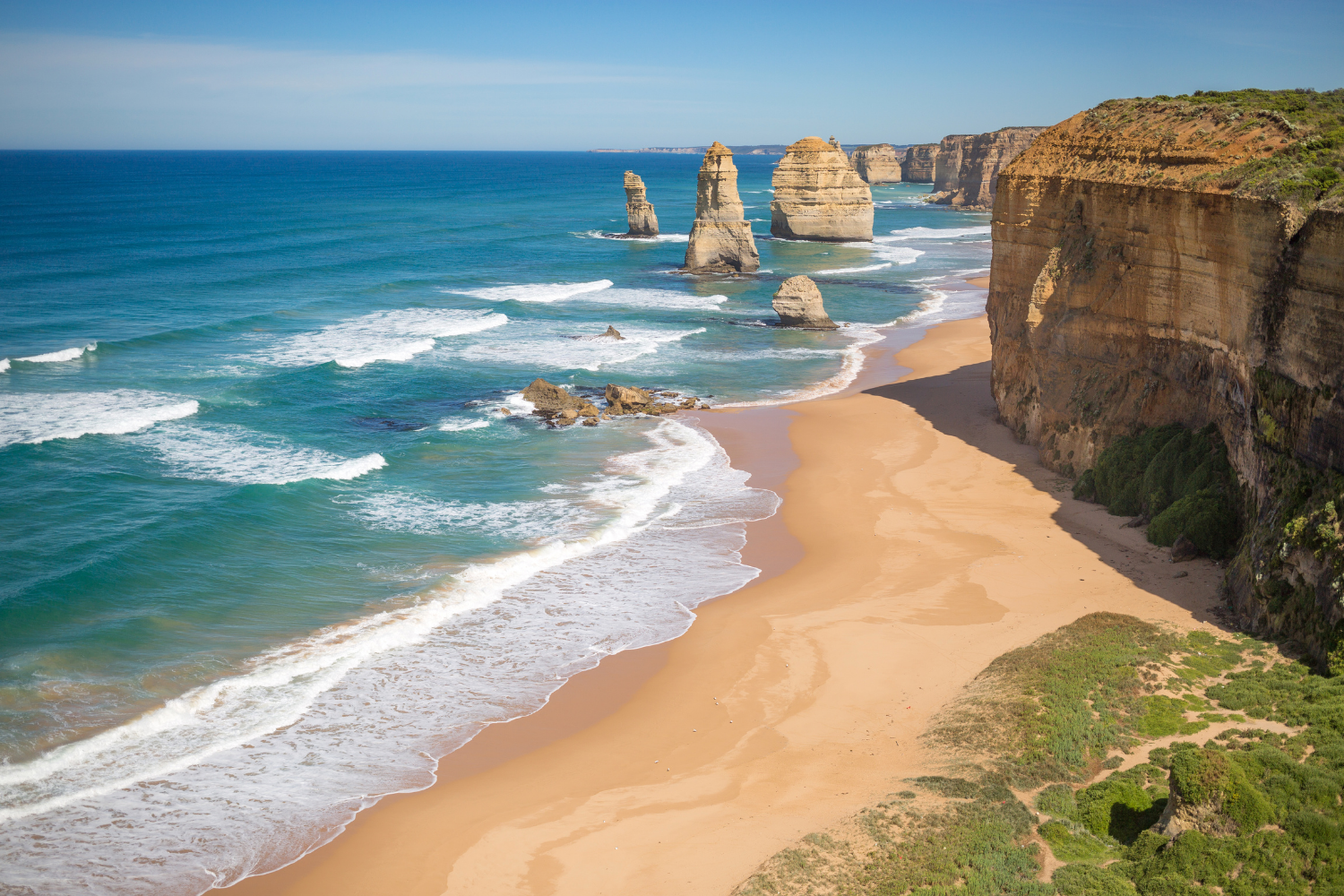A scenic view of the Twelve Apostles limestone stacks along the southern coast of Australia, with turquoise ocean waves crashing onto a sandy beach and rugged cliffs topped with green vegetation.