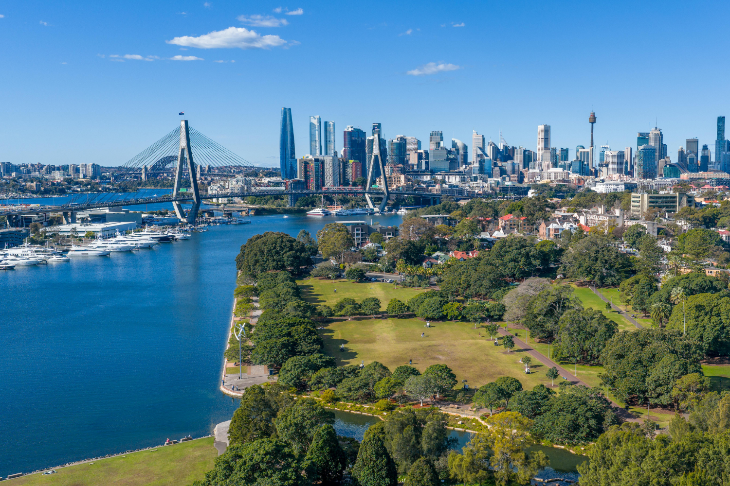 Aerial view of a city skyline with a river, bridge, and parks in the foreground, featuring tall modern buildings and clear blue sky.