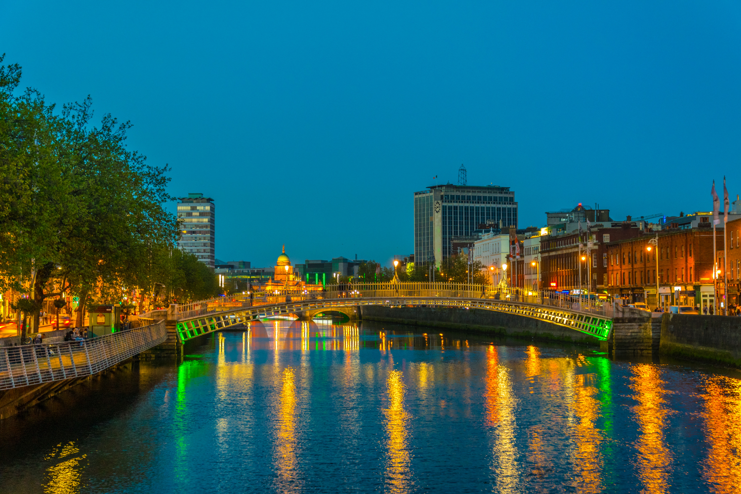 Cityscape at dusk with a river in the foreground reflecting colorful lights, a bridge, historic buildings, and modern skyscrapers lining the waterfront.