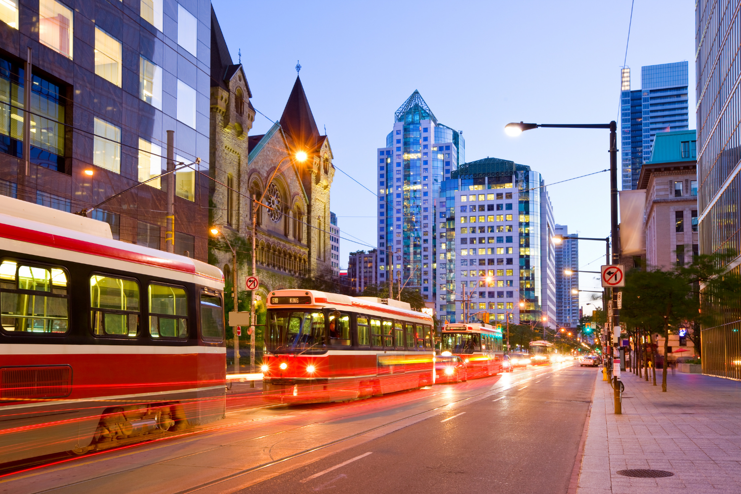 City street scene at dusk with multiple light rail vehicles moving along tracks, tall modern skyscrapers, and historic church building, illuminated streetlights, and light trails from vehicle motion.