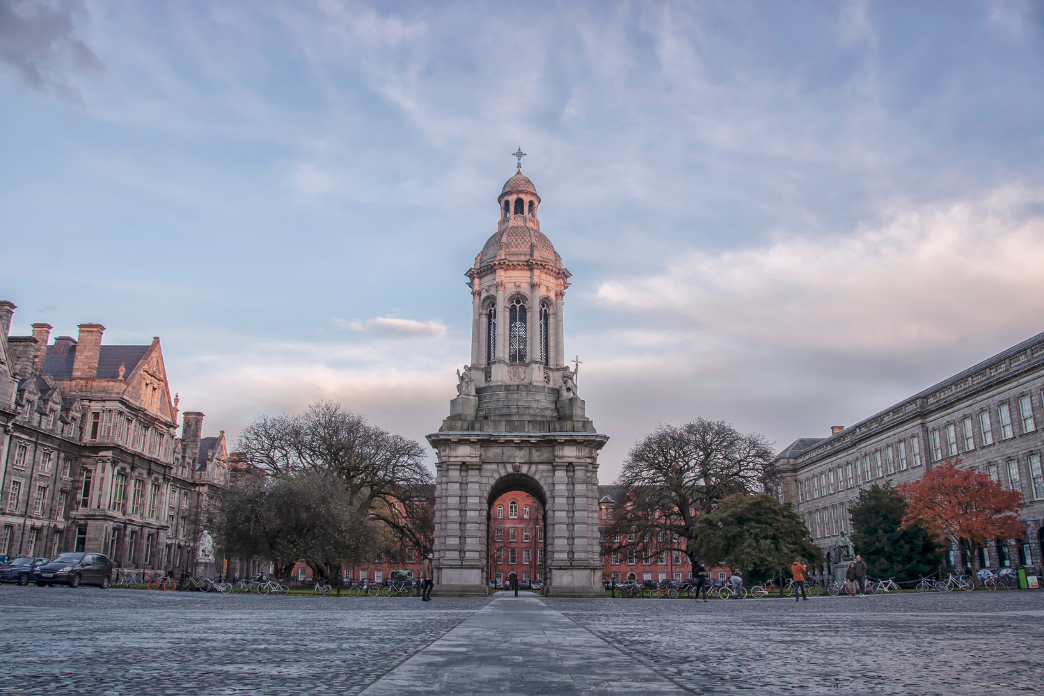 A historic clock tower with an archway at its base, situated in the middle of a cobblestone square with trees and buildings around, during sunset with a partly cloudy sky.