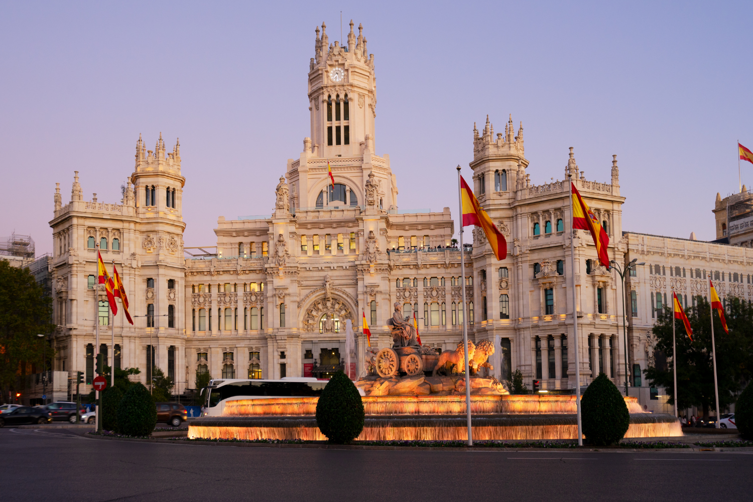 Historic white building with a clock tower, flags, and a fountain in front at sunset, resembling a government or royal building.