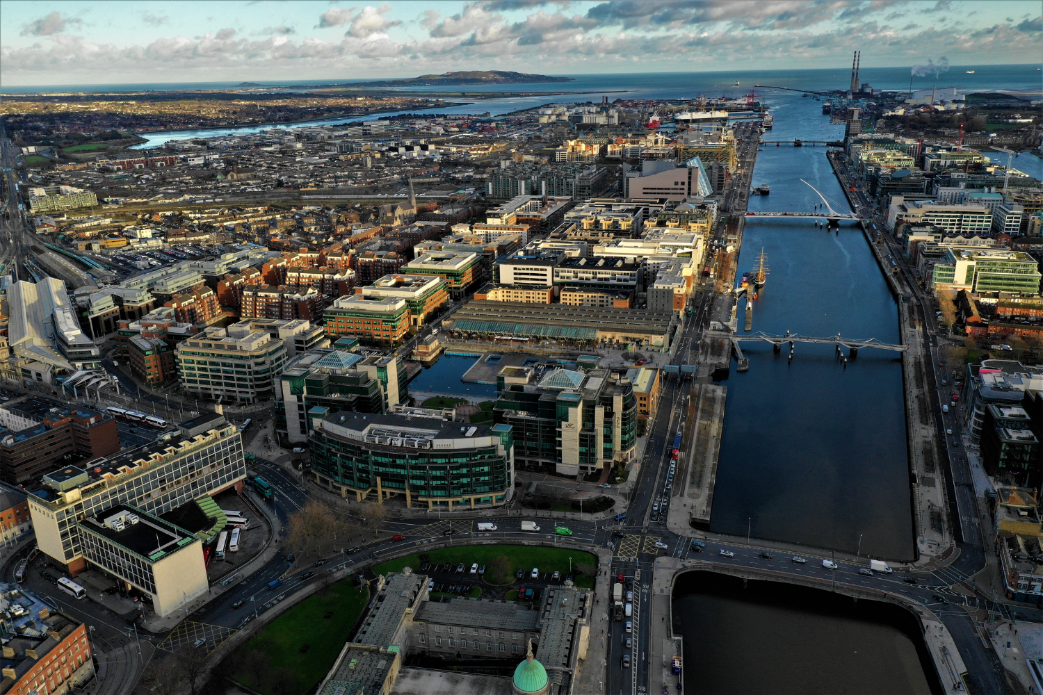 Aerial view of a city with modern buildings, waterways, and bridges, with a harbor and distant hills under a partly cloudy sky.