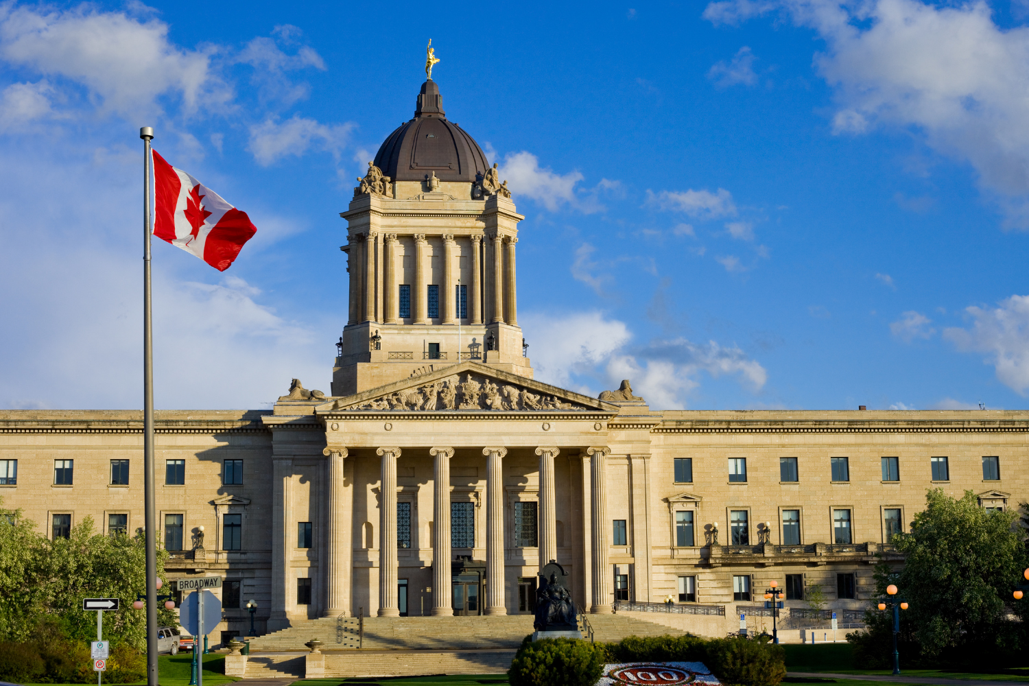 The Historic City Hall in Kingston, Ontario, Canada, with a Canadian flag flying on a pole in the foreground, a statue in front, and a blue sky with scattered clouds overhead.