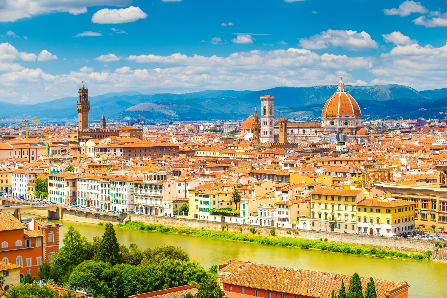 Cityscape of Florence, Italy, featuring historic buildings, the Florence Cathedral with its large dome, the Palazzo Vecchio tower, and a river running through the city with mountains in the background.