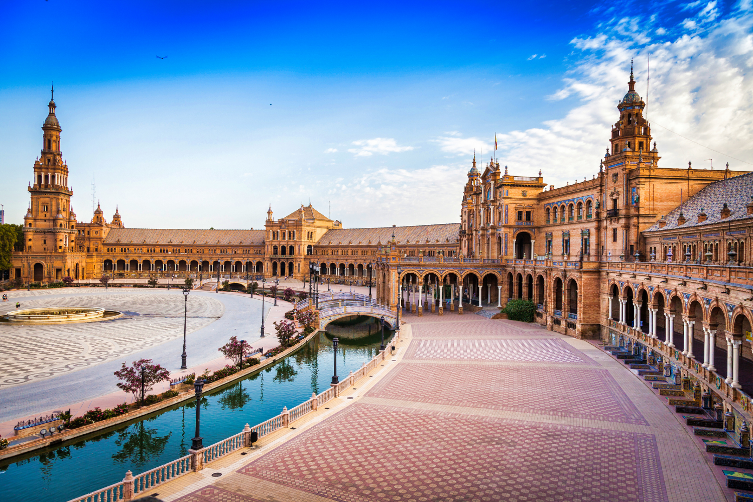 Historic building with tall towers, arches, and a courtyard featuring a fountain, trees, and a canal, under a partly cloudy sky.
