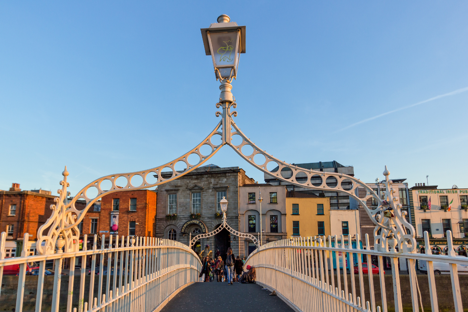 Pedestrian bridge with decorative white metal arches and street lamps, leading to historic buildings and a pub, under a clear blue sky during sunset.