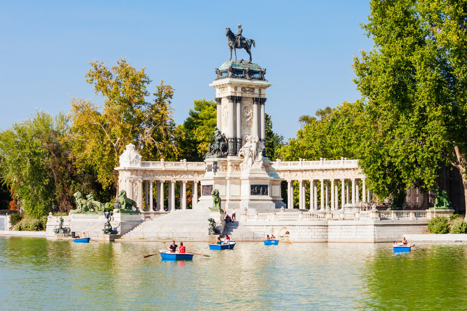 People rowing boats in a body of water with a large ornate monument featuring statues and a horse statue on top, surrounded by trees.