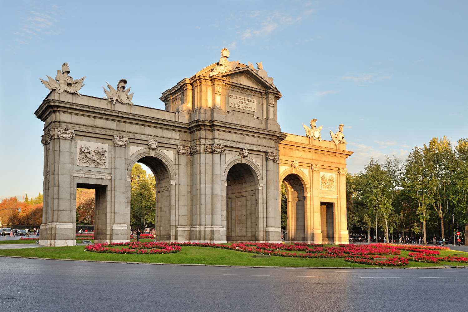 A stone arch monument with decorative sculptures and inscriptions illuminated by sunset, surrounded by flower beds and trees, with a clear blue sky in the background.