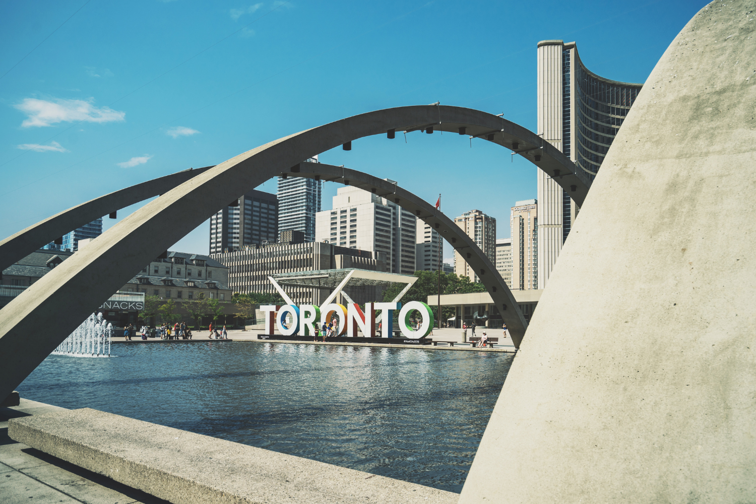 Cityscape of Toronto with a large colorful 'TORONTO' sign near a fountain and a reflective pool, framed by modern concrete arches and tall skyscrapers under a blue sky.