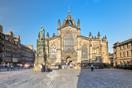 A large historic church with Gothic architecture featuring pointed arches and tall spires, surrounded by a cobblestone square with statues and people walking around.