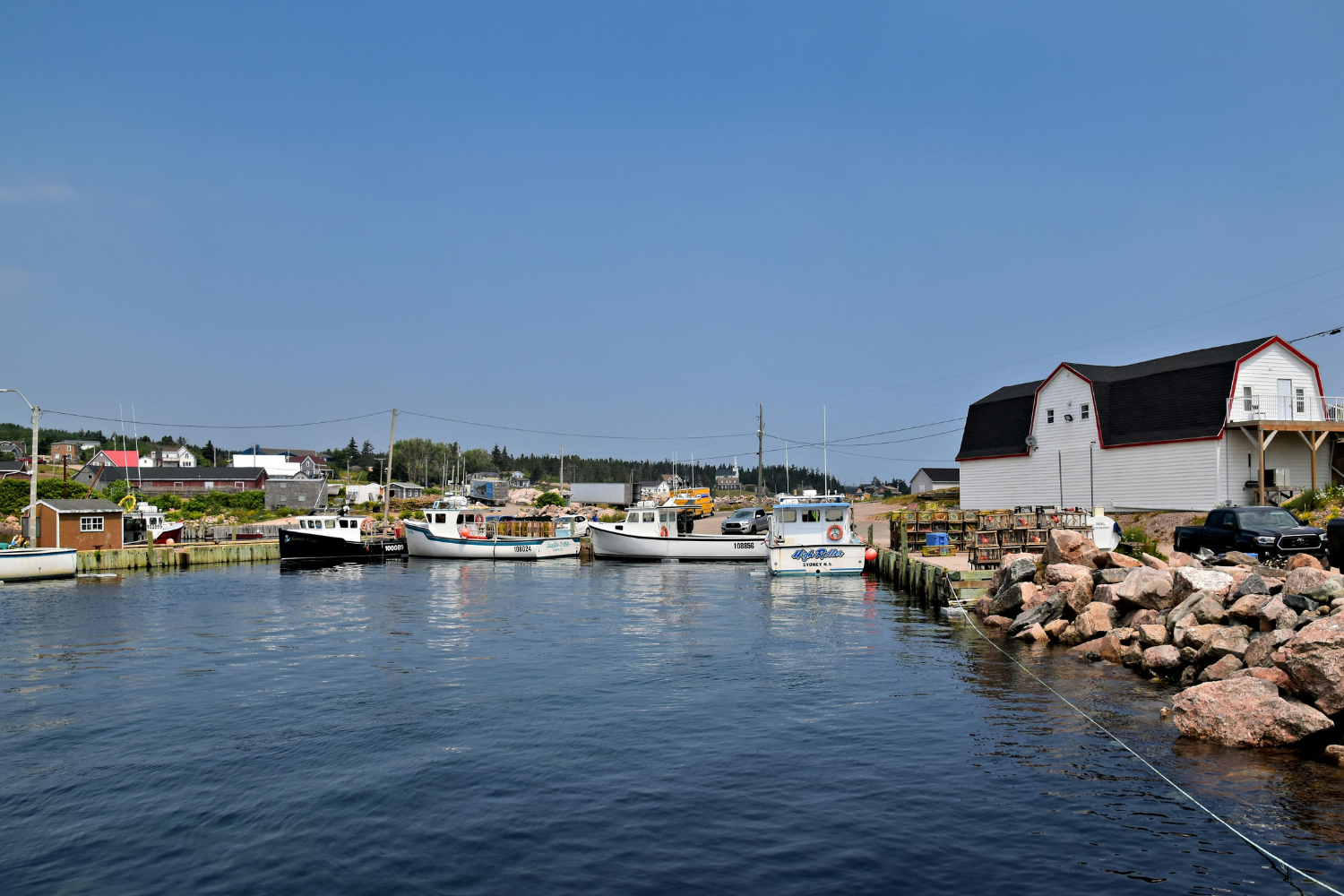 A marina with four boats docked along the water, a large white barn with a black roof and red trim, and cars parked nearby. Rocks line the shoreline, and houses and trees are in the background under a clear blue sky.