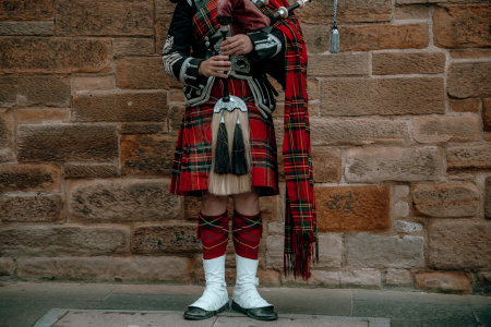 Person wearing traditional Scottish attire playing bagpipes in front of a stone wall.