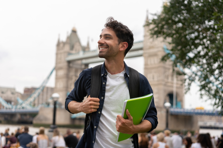 Man holding a green notebook, smiling, near Tower Bridge in London, with tourists and trees in the background.
