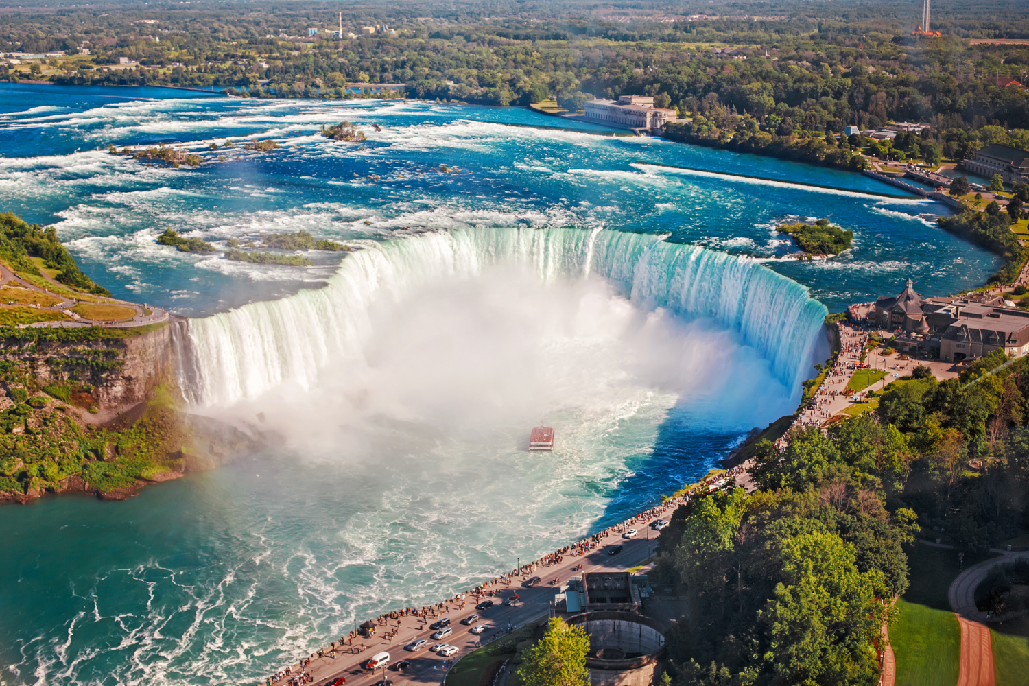 Aerial view of the waterfalls at Niagara Falls with a boat near the base, crowds of tourists along the walkway, and lush green surroundings.
