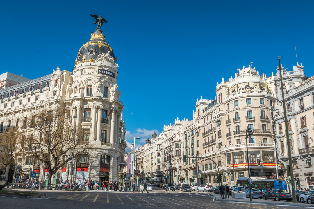 City street scene with historic white buildings, a prominent building with a copper dome and a statue of a winged figure on top, and a blue sky in Madrid, Spain.