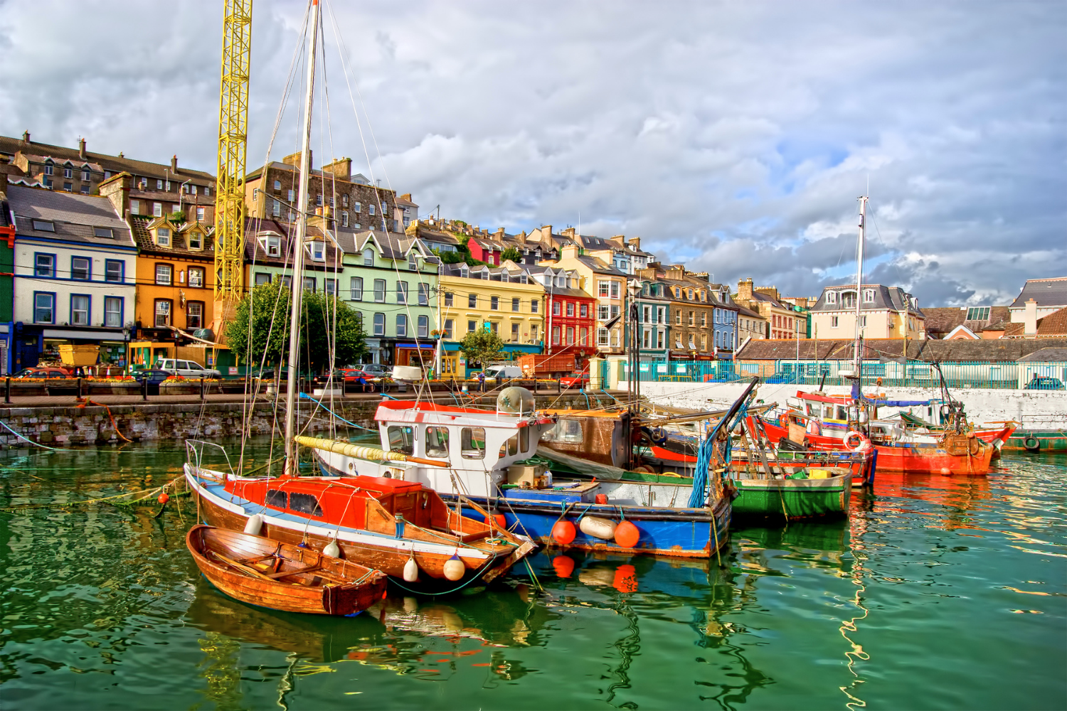 Boats docked at a colorful harbor with a row of multi-colored houses along the waterfront, under a cloudy sky.