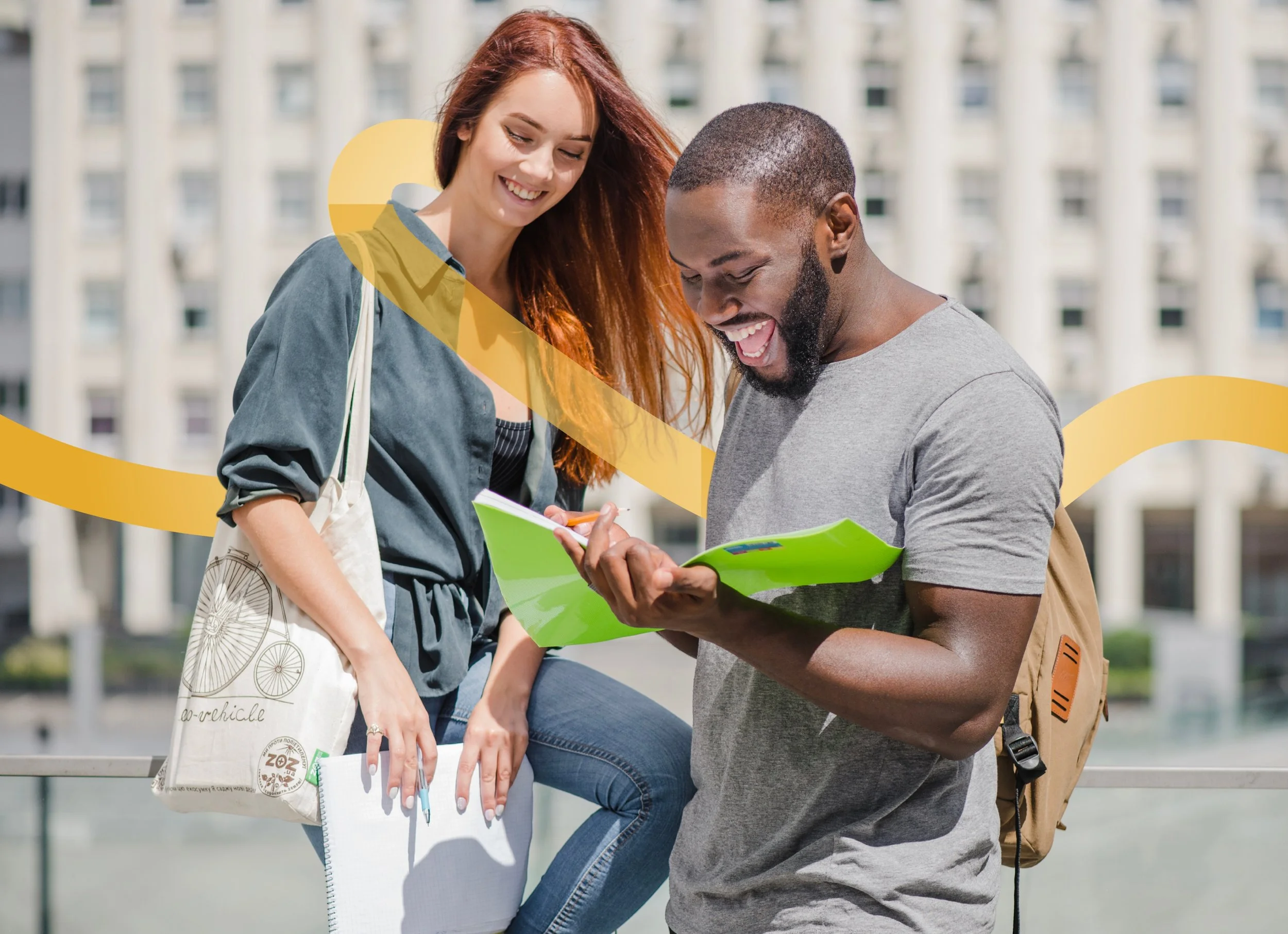 Two young adults, a woman with long red hair and a man with a beard, are looking at a notebook and smiling. The woman is carrying a tote bag and a journal, and the man is wearing a backpack and holding a green folder. They are outdoors in an urban setting with a large building in the background.