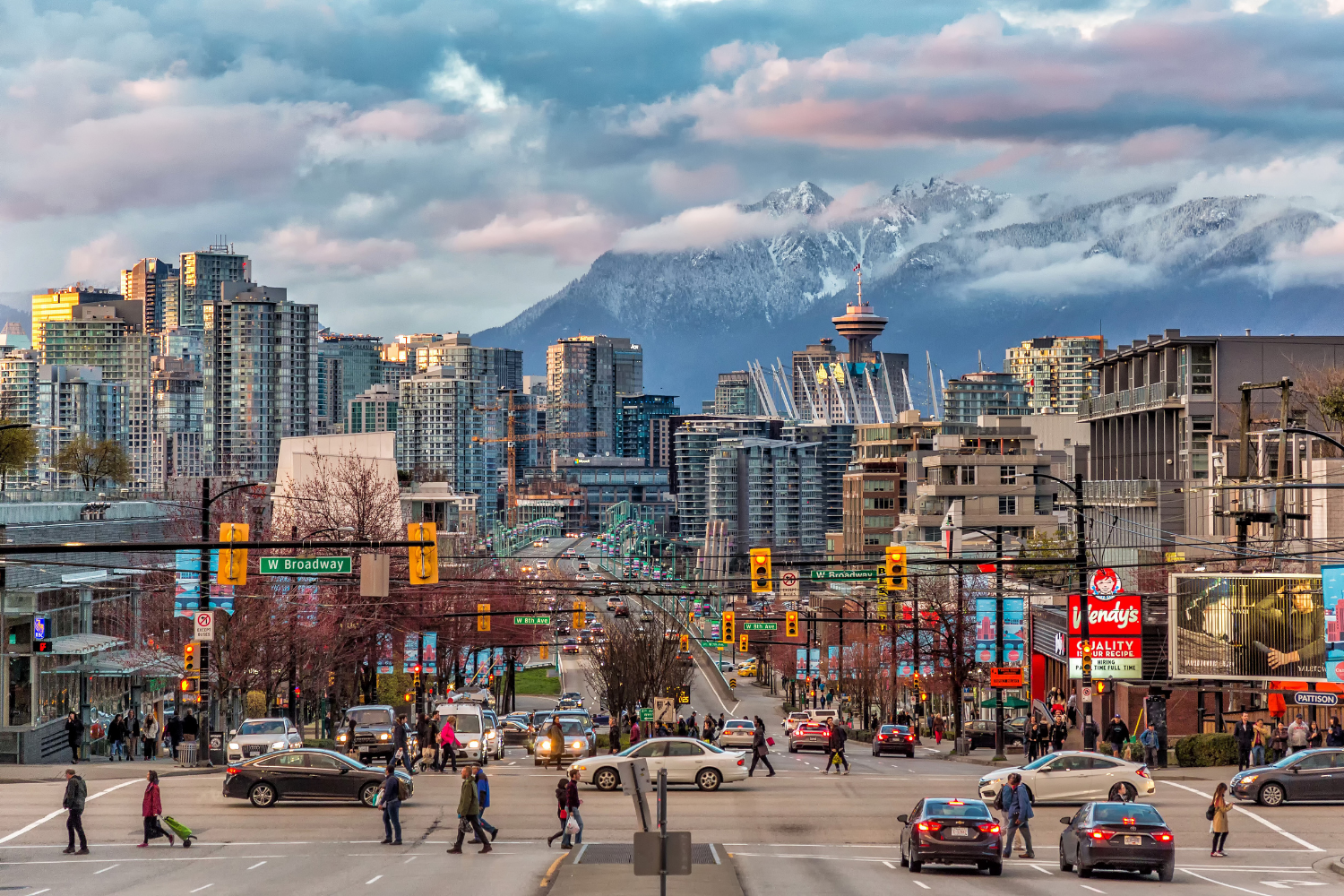 Urban city street with pedestrians crossing, cars on the road, and high-rise buildings with mountain range in the background, with Mount Rainier visible.