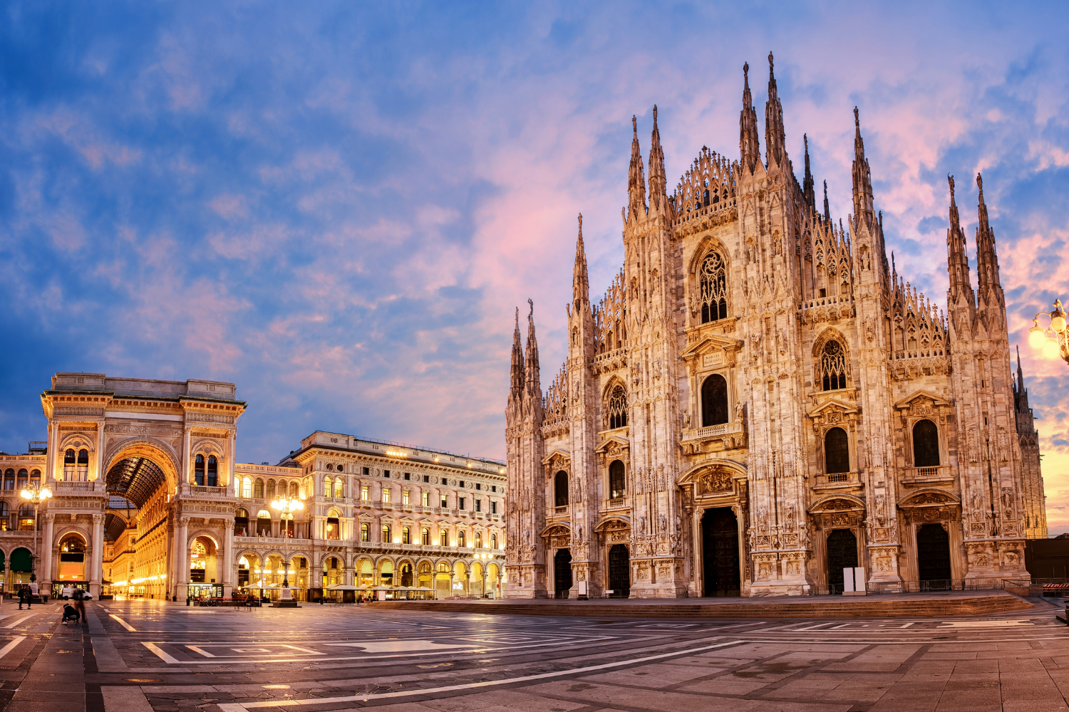 The Milan Cathedral (Duomo di Milano), a large Gothic cathedral with intricate marble facade and spires, during sunset in Piazza del Duomo with illuminated surrounding buildings and a colorful sky.