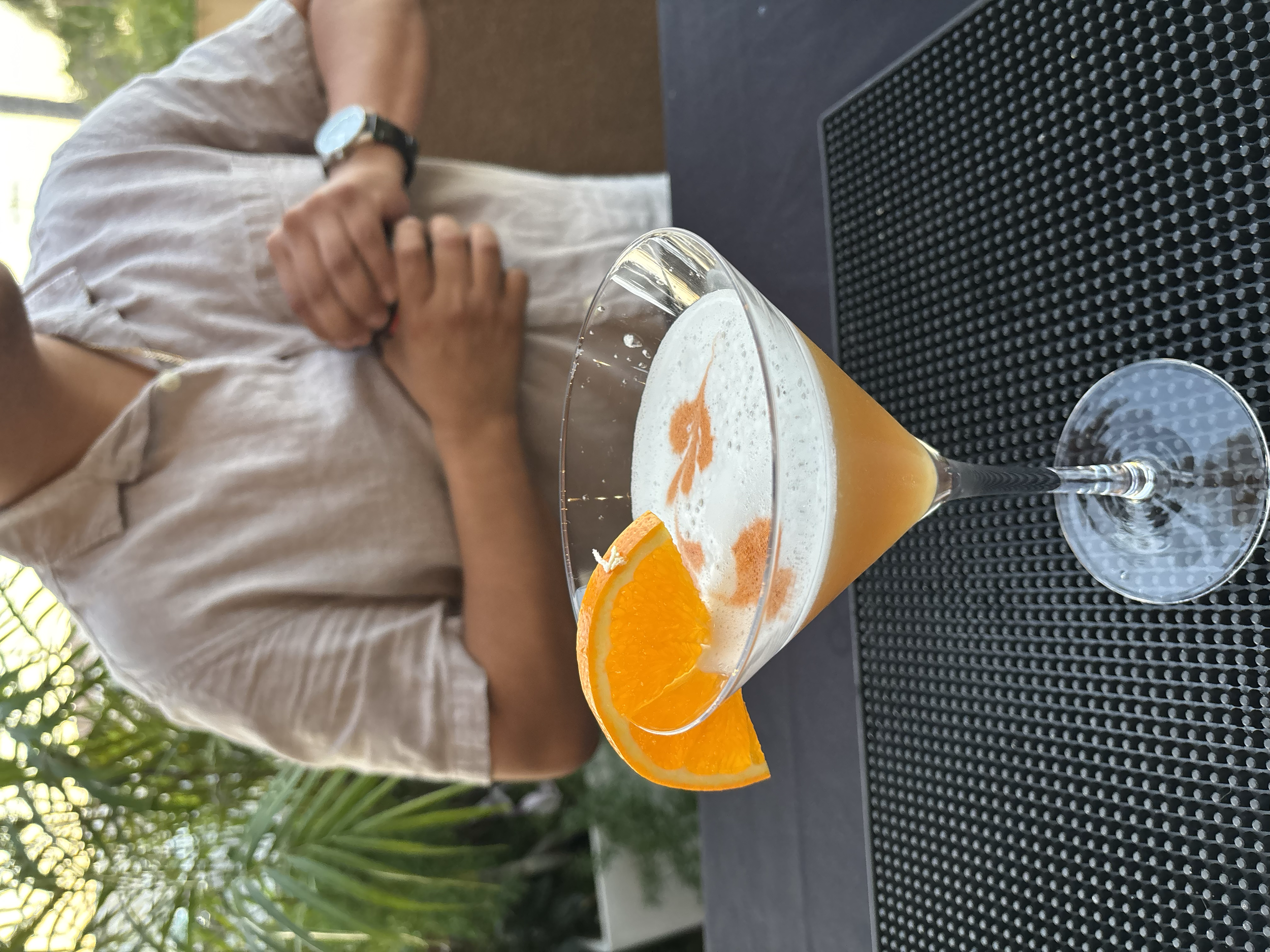 Close-up of a cocktail glass filled with an orange-colored drink, garnished with an orange wedge. A person wearing a beige shirt and a watch is sitting at a table, with their hands clasped, in the background.