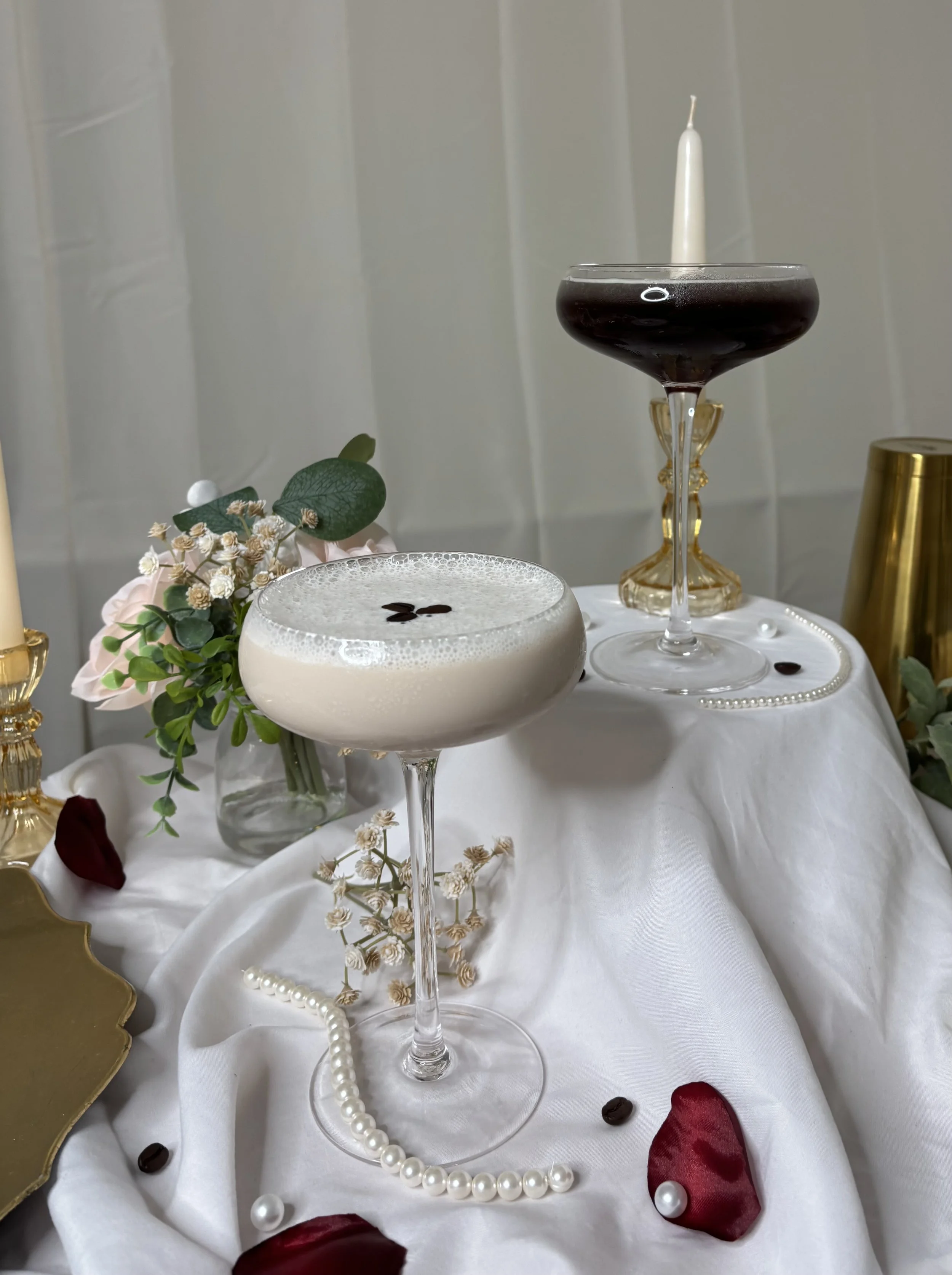 Elegant table arrangement featuring two cocktails, one with foam and coffee beans, and the other dark and layered, on a white cloth with candles, flowers, pearls, and rose petals.
