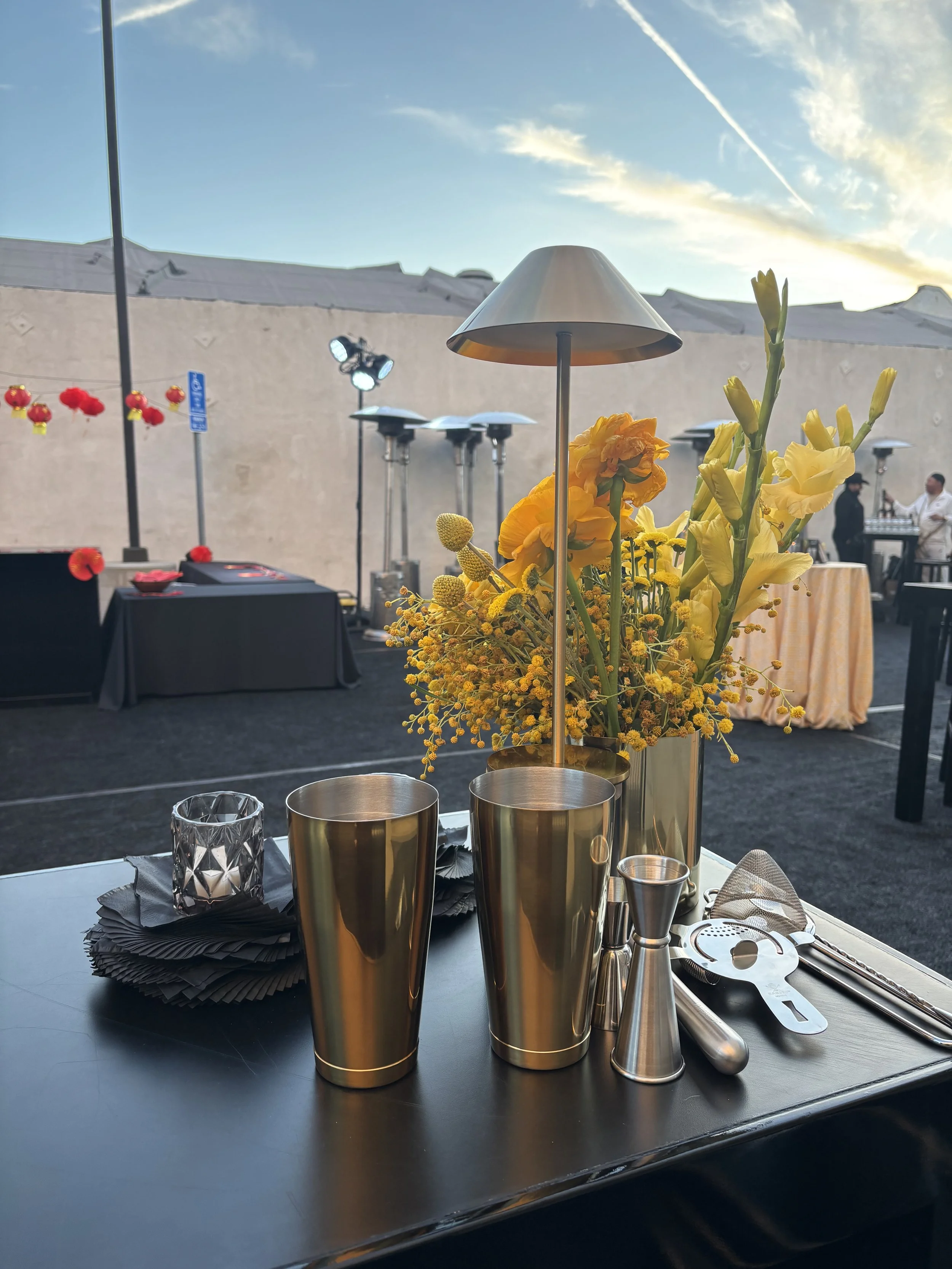 A table set with a modern floral centerpiece and metallic cocktail shakers at an outdoor event during sunset, with lighting equipment and a DJ in the background.