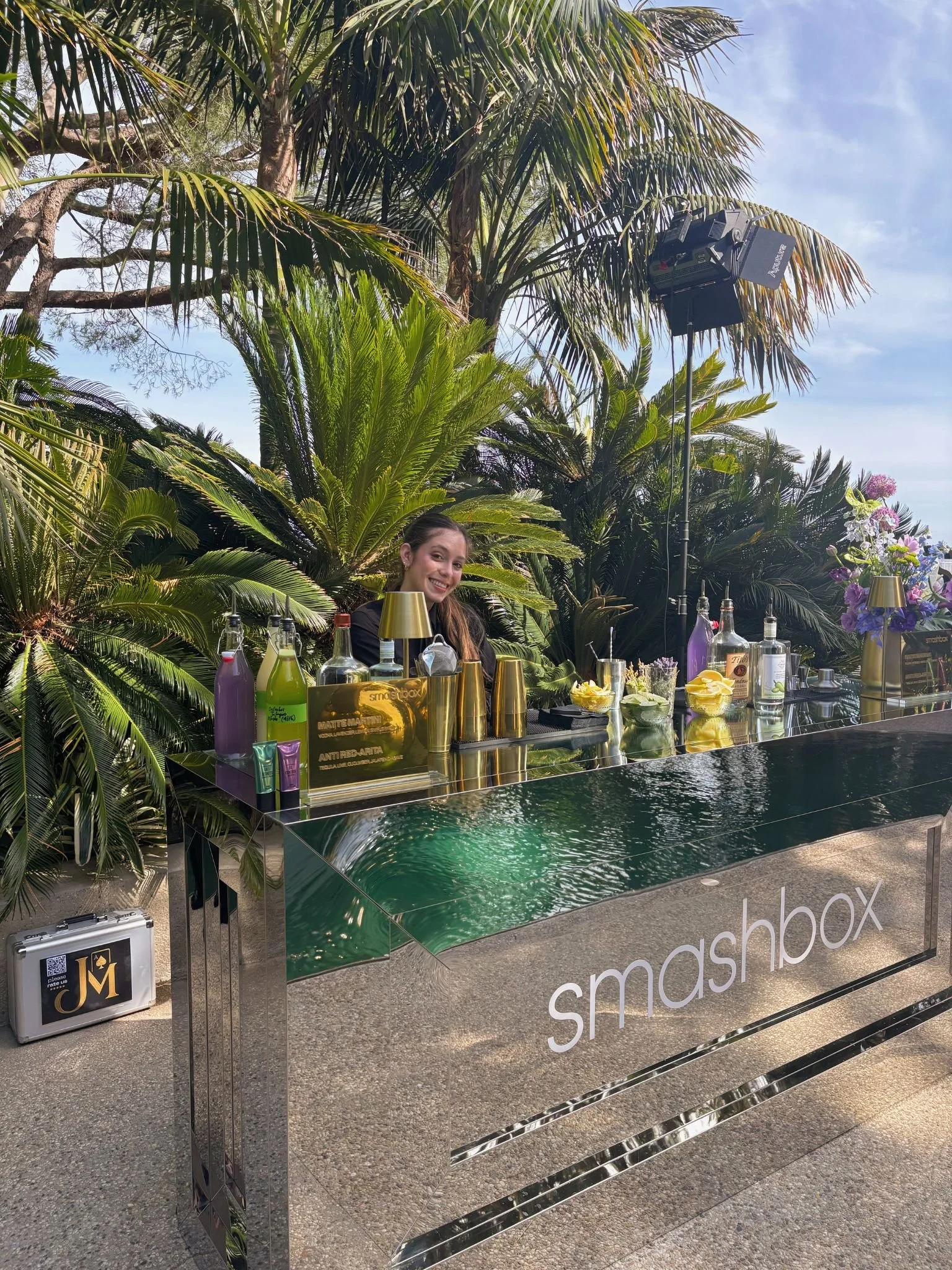A woman smiling behind a reflective bar with the word 'smashbox' on it, surrounded by lush green tropical plants, with a clear blue sky and palm trees in the background.