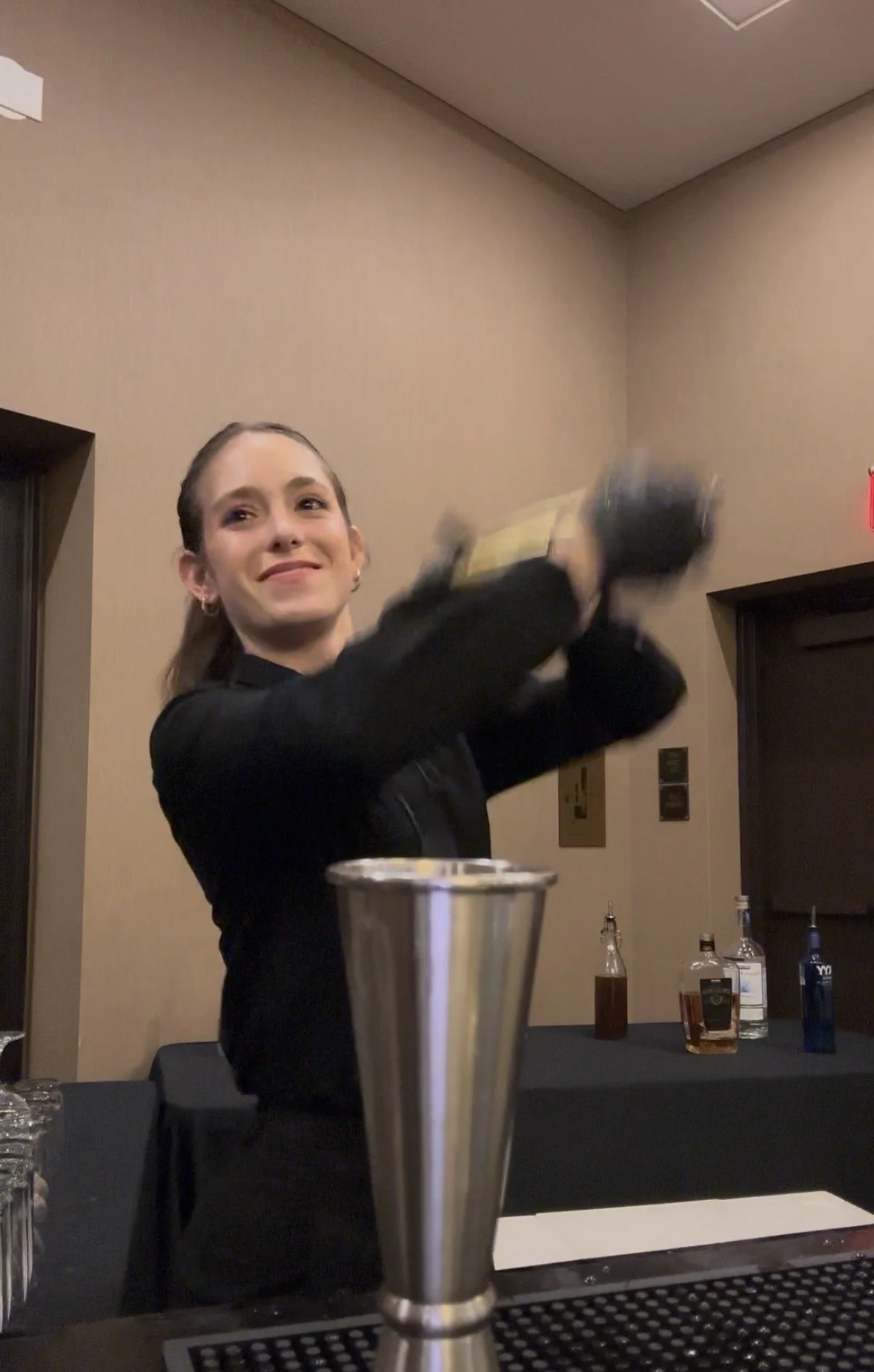 A young woman mixing a drink at a bar, smiling, with bottles of liquor in the background.