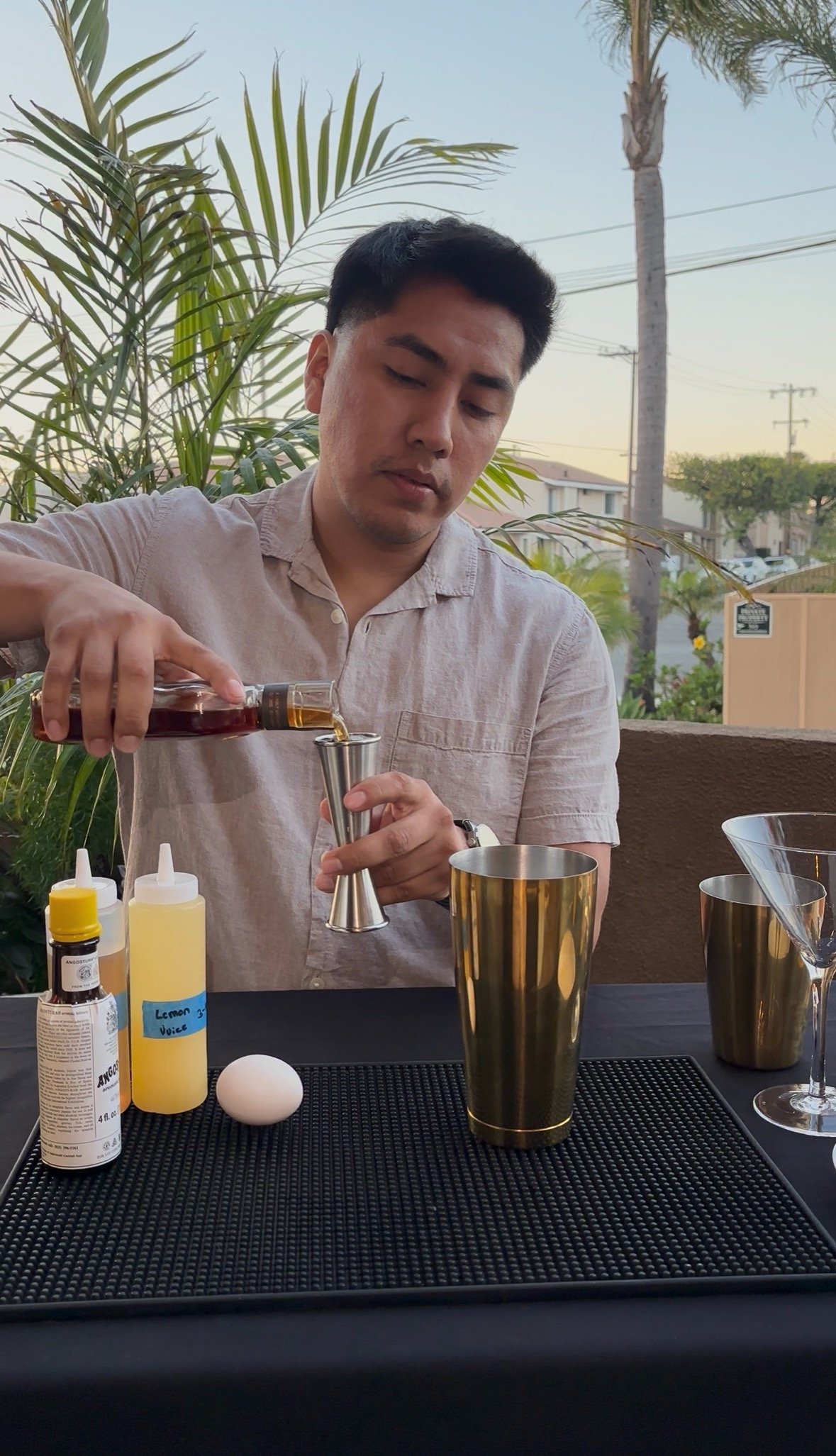 A man preparing a cocktail outdoors at sunset, with various bar tools and ingredients on the table, including a bottle of liquor, lemon juice, and an egg.