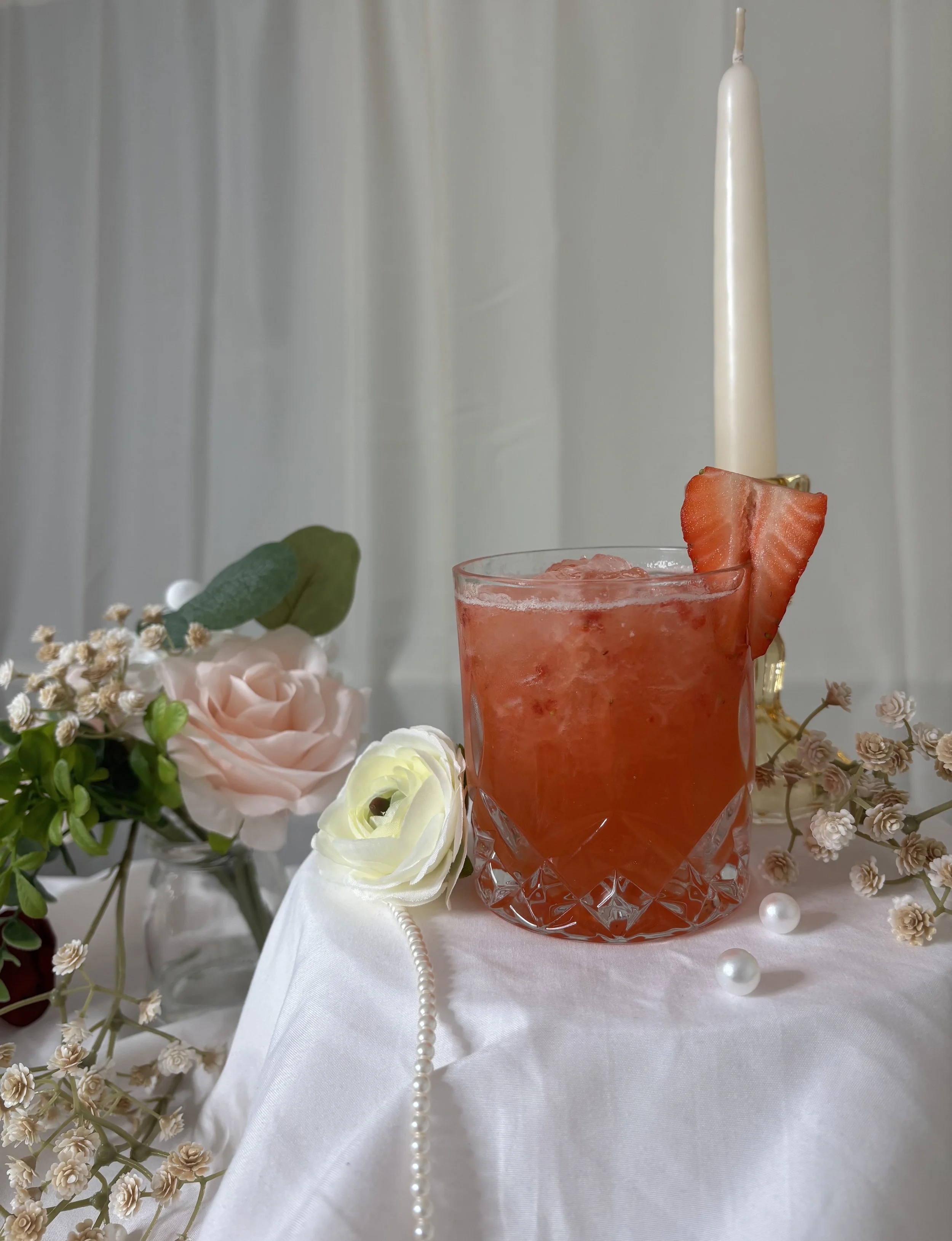 A glass of pink strawberry cocktail garnished with a slice of strawberry, surrounded by flowers and pearls on a white tablecloth, with a white candle in the background.