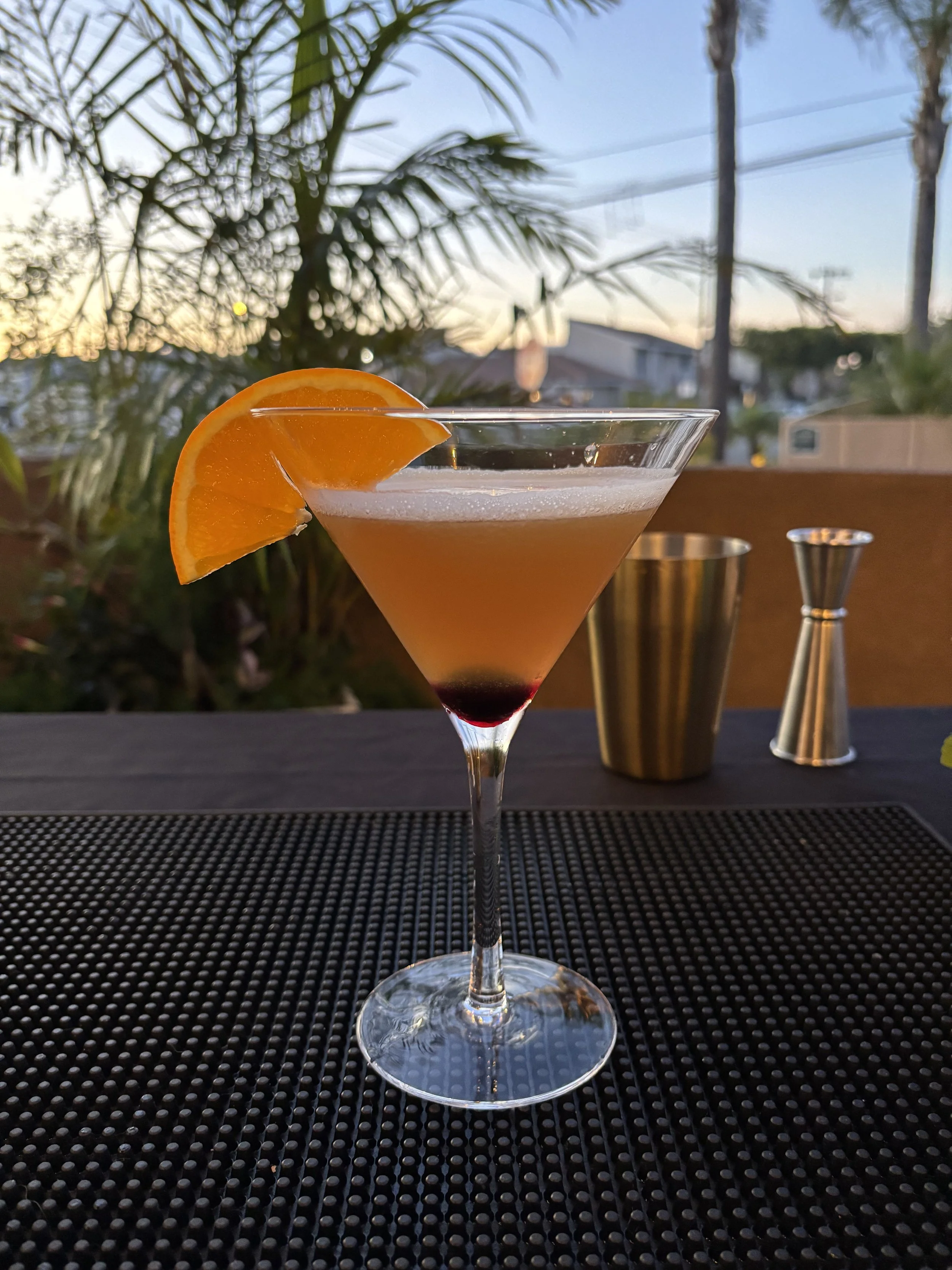 A cocktail glass with an orange slice garnish sitting on a black textured placemat outdoors during sunset, with a blurred background of plants, houses, and a sky transitioning from blue to orange.