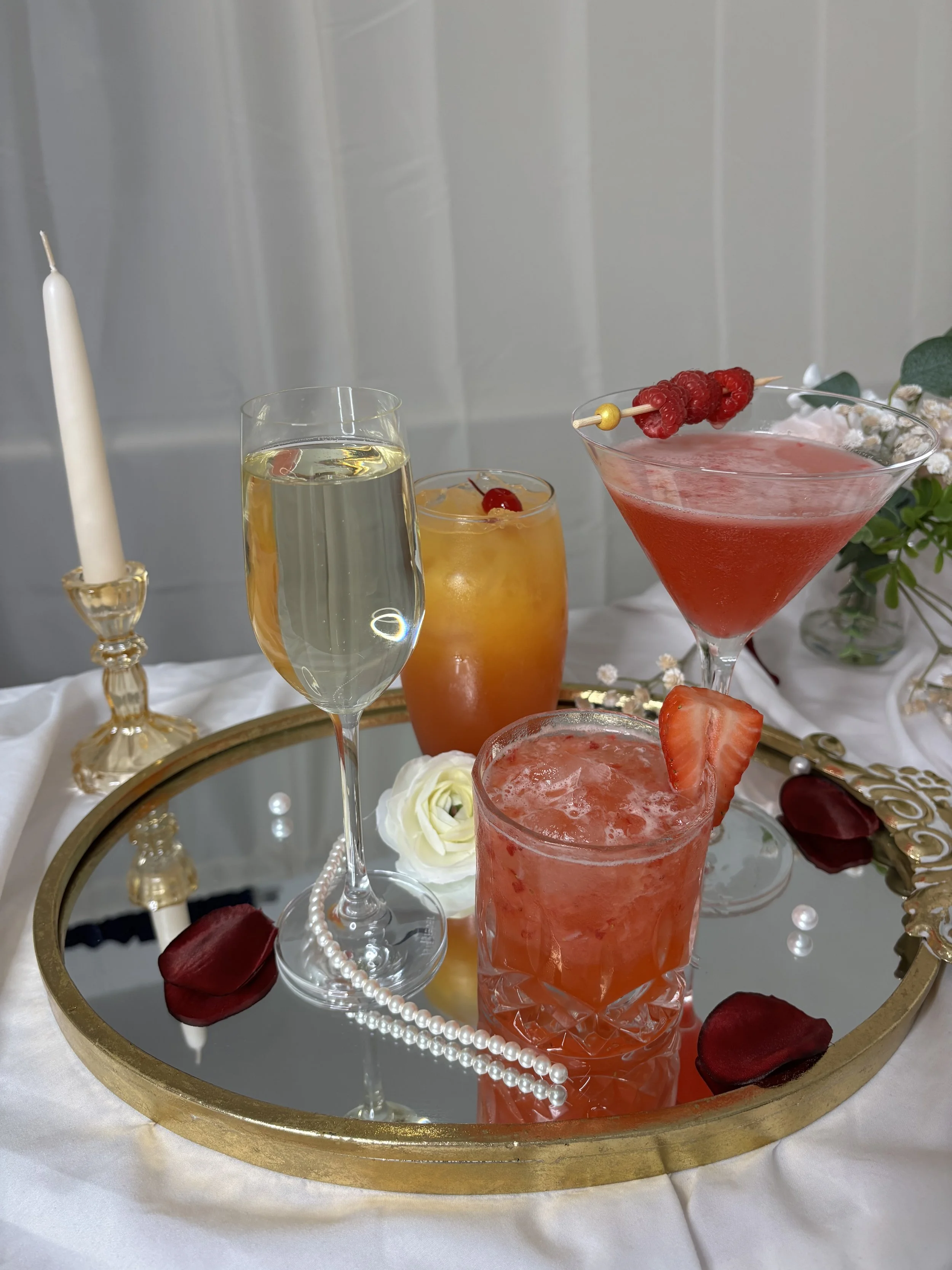 A decorative display of five cocktails and beverages on a gold-framed mirror tray, surrounded by rose petals, a string of pearls, a white rose, and a candle, with a white tablecloth and floral arrangement in the background.
